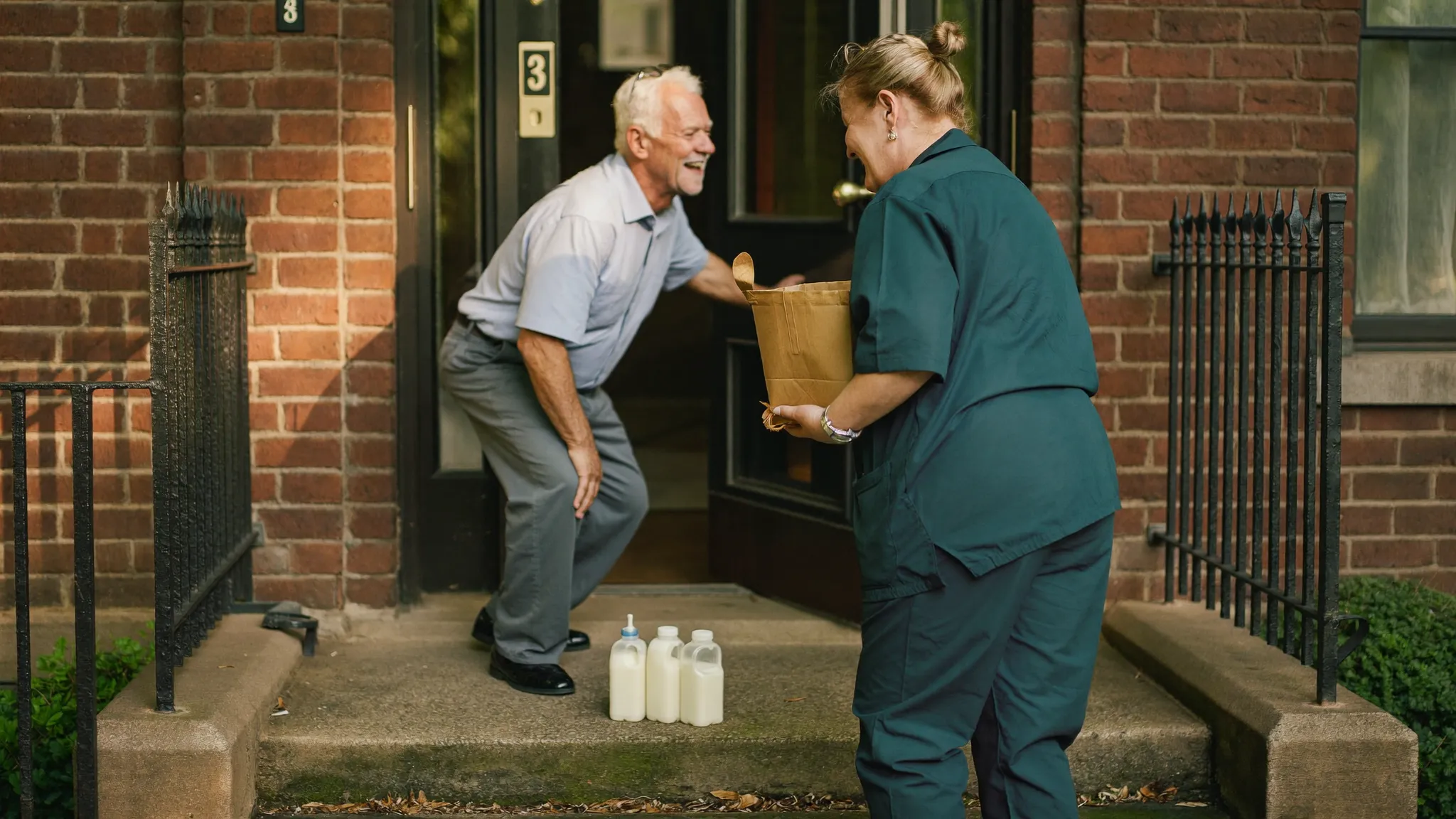 A care worker in a green tunic laughing with an older man as he opens the front door of a red-brick terraced house in the morning