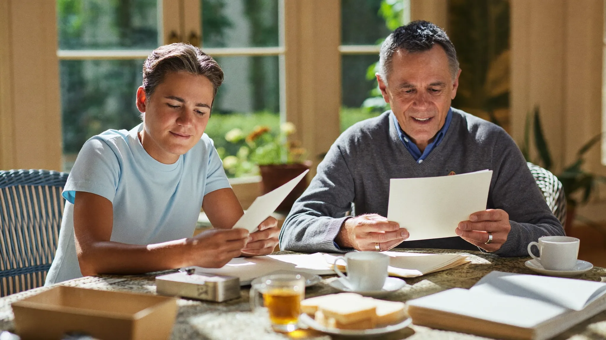 An adult son and his father sorting official letters together at a conservatory table with mugs of tea and a biscuit tin