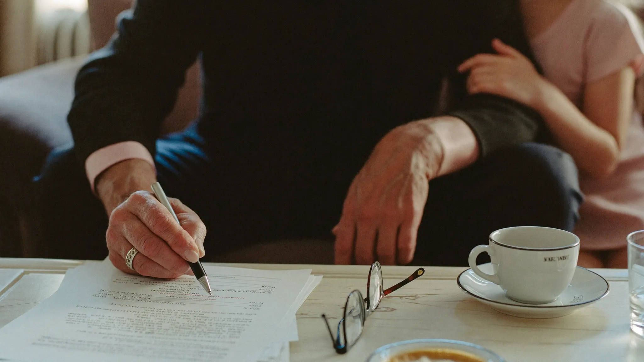 An older man's hands holding a pen over forms on a coffee table, reading glasses and tea beside him, with his daughter's hand resting on his shoulder