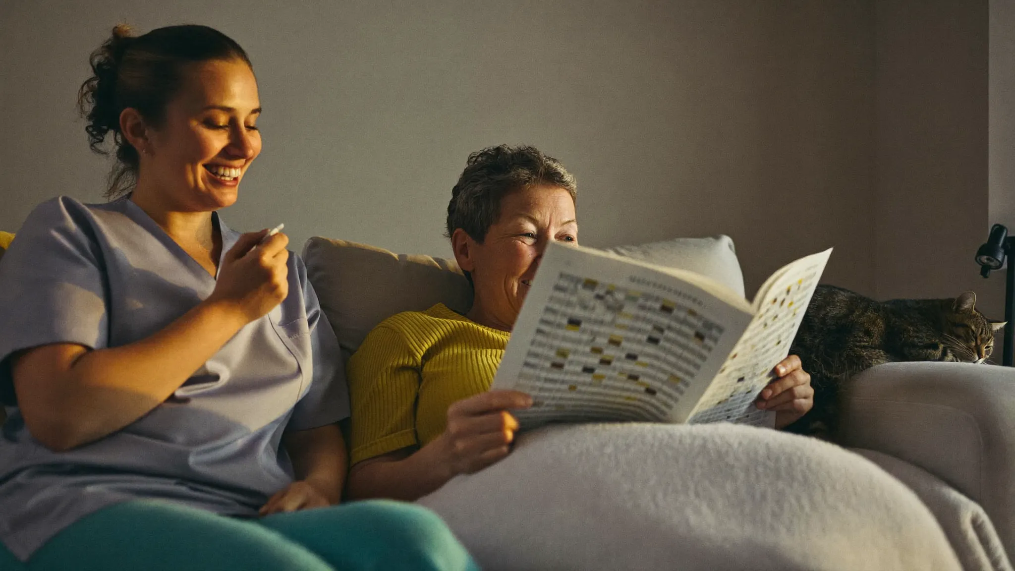 A live-in carer in a green tunic and an older woman laughing over a crossword on the sofa, a cat asleep on the armchair