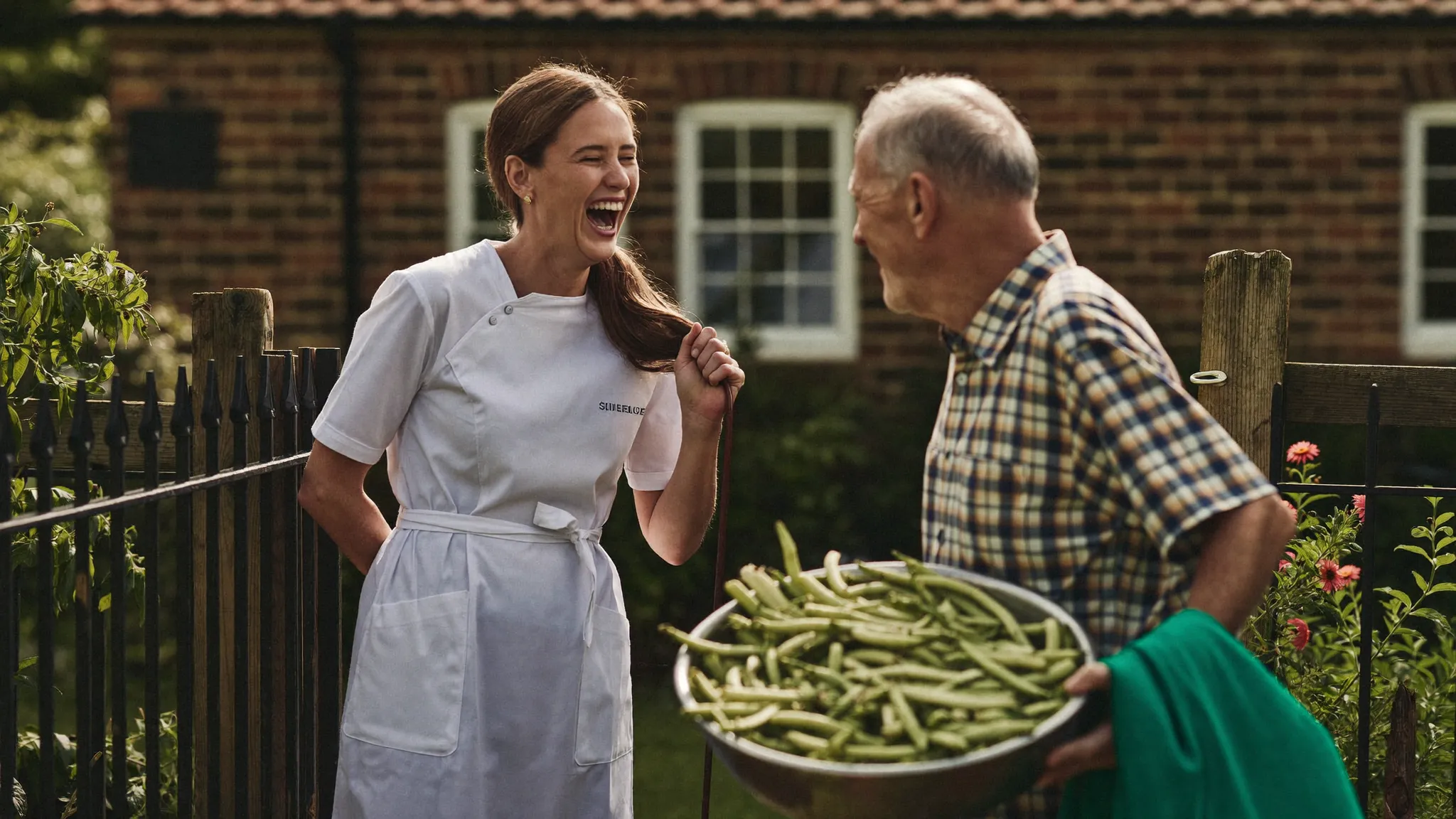 A care worker in a green tunic laughing with an older man holding a trug of runner beans by his garden gate