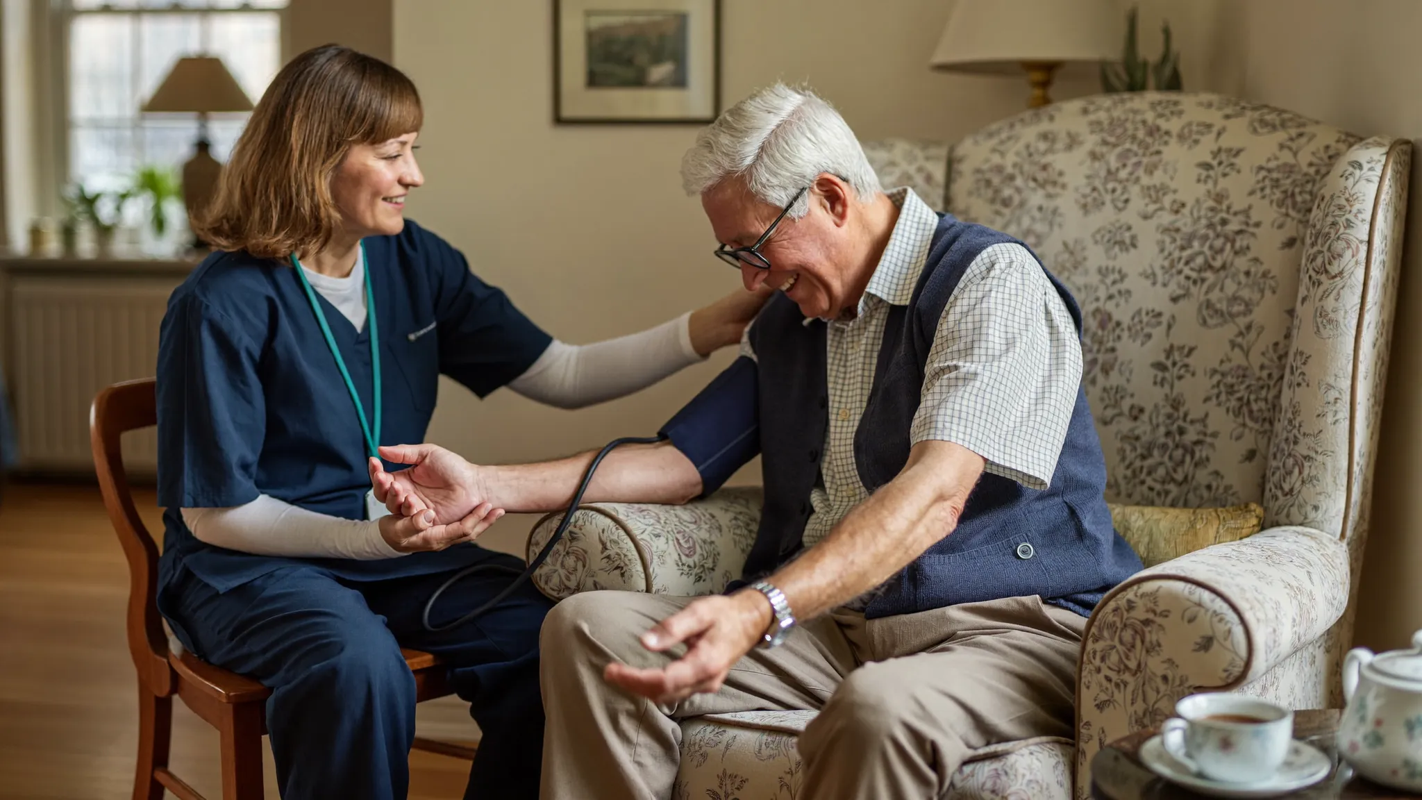 A community nurse in a navy tunic checking the blood pressure of a smiling older man seated in a floral armchair at home